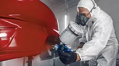 Man spraying finishing layer of paint to vehicle part. 