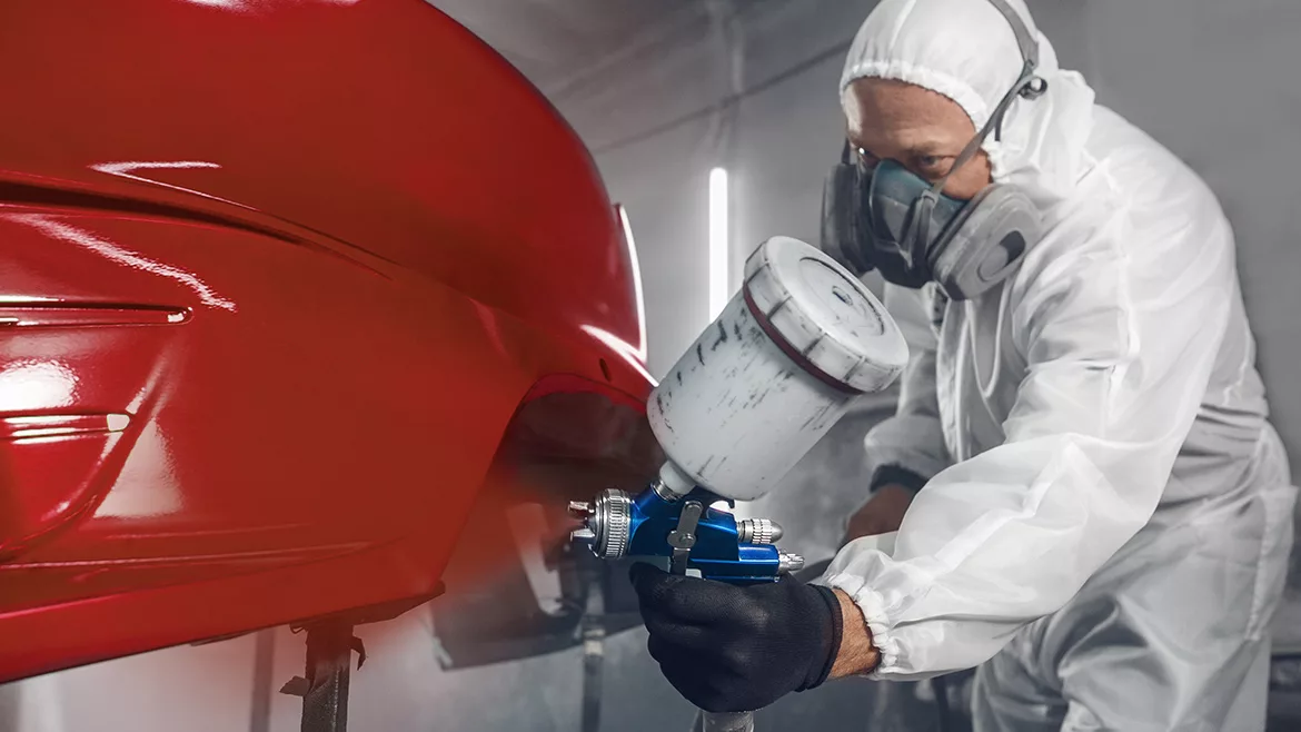 Man spraying finishing layer of paint to vehicle part.