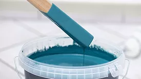 Paintbrush dipped in vibrant teal paint, hovering above a bucket, showcasing the artistic process and creativity in home improvement projects
