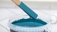 Paintbrush dipped in vibrant teal paint, hovering above a bucket, showcasing the artistic process and creativity in home improvement projects