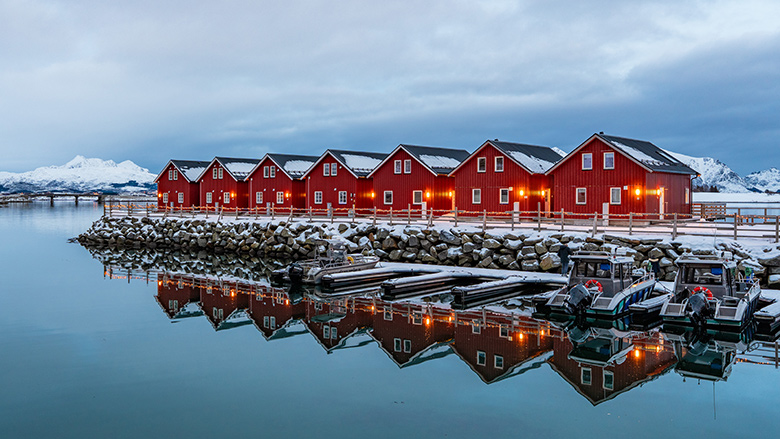Red rorbuer cabins line a snowy harbor reflecting in still water with mountains