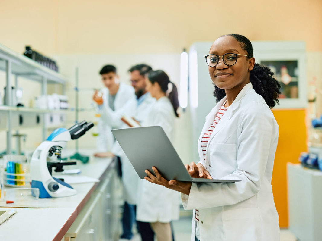 Happy black female chemist working on laptop in laboratory and looking at camera.